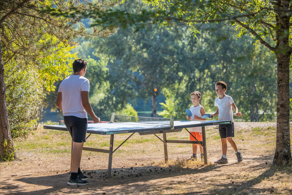 Partie de ping-pong: trois enfants jouent à l'ombre des arbres du parc des Chalets de Booz. Dans le fond, on voit les arbres en bord de rivière