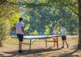 Partie de ping-pong: trois enfants jouent à l'ombre des arbres du parc des Chalets de Booz. Dans le fond, on voit les arbres en bord de rivière
